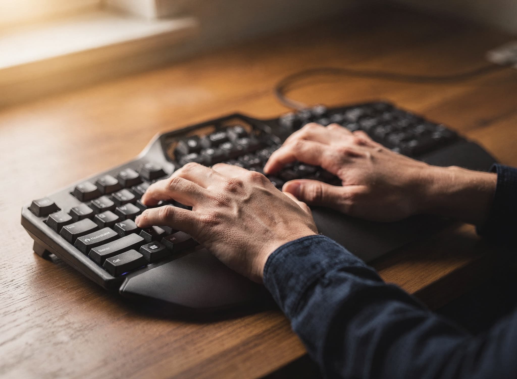 Mani che digitano su una tastiera ergonomica divisa nera con poggiapolsi integrati su una calda scrivania in legno.